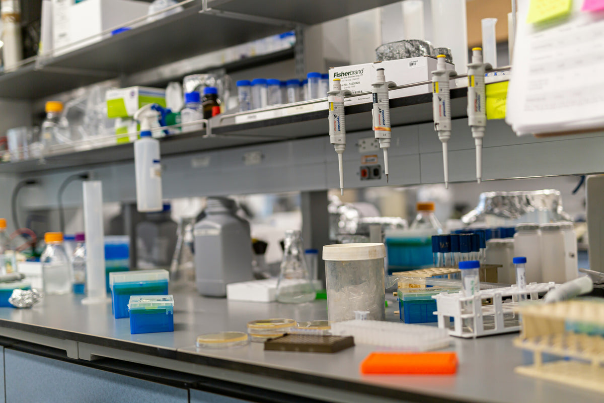 A laboratory workbench with multiple pipettes hanging from a rail, test tubes, bottles, petri dishes, and other scientific equipment in a real indoor setting. | Labs Choice